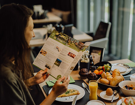 Eine Frau liest während des Frühstücks im Lürzerhof Gourmet & Wellnesshotel eine Broschüre mit Wellness- und Freizeitangeboten.
